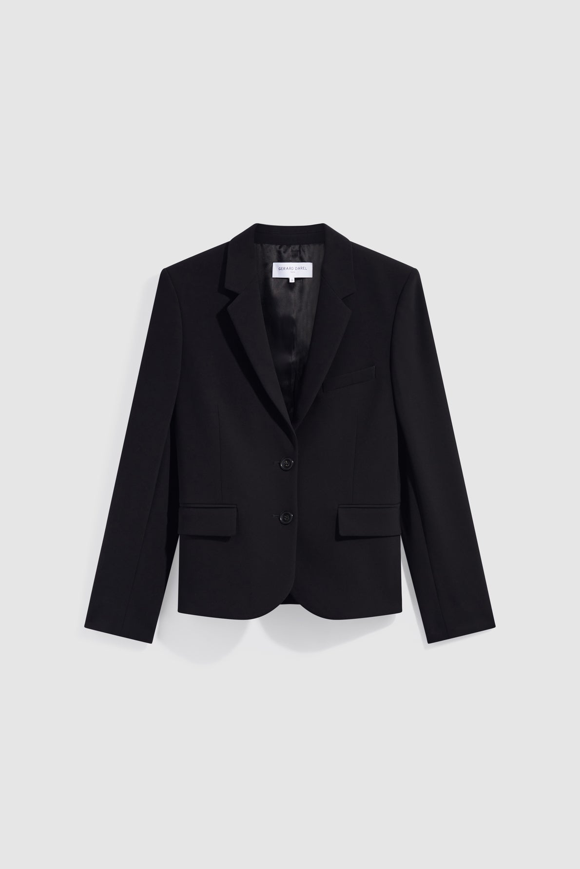 A black tailored blazer with notch lapels, two front buttons, flap pockets, and a chest pocket is displayed against a plain light gray background. This veste noire de tailleur exudes a chic sophistication, perfect for elevating any ensemble.