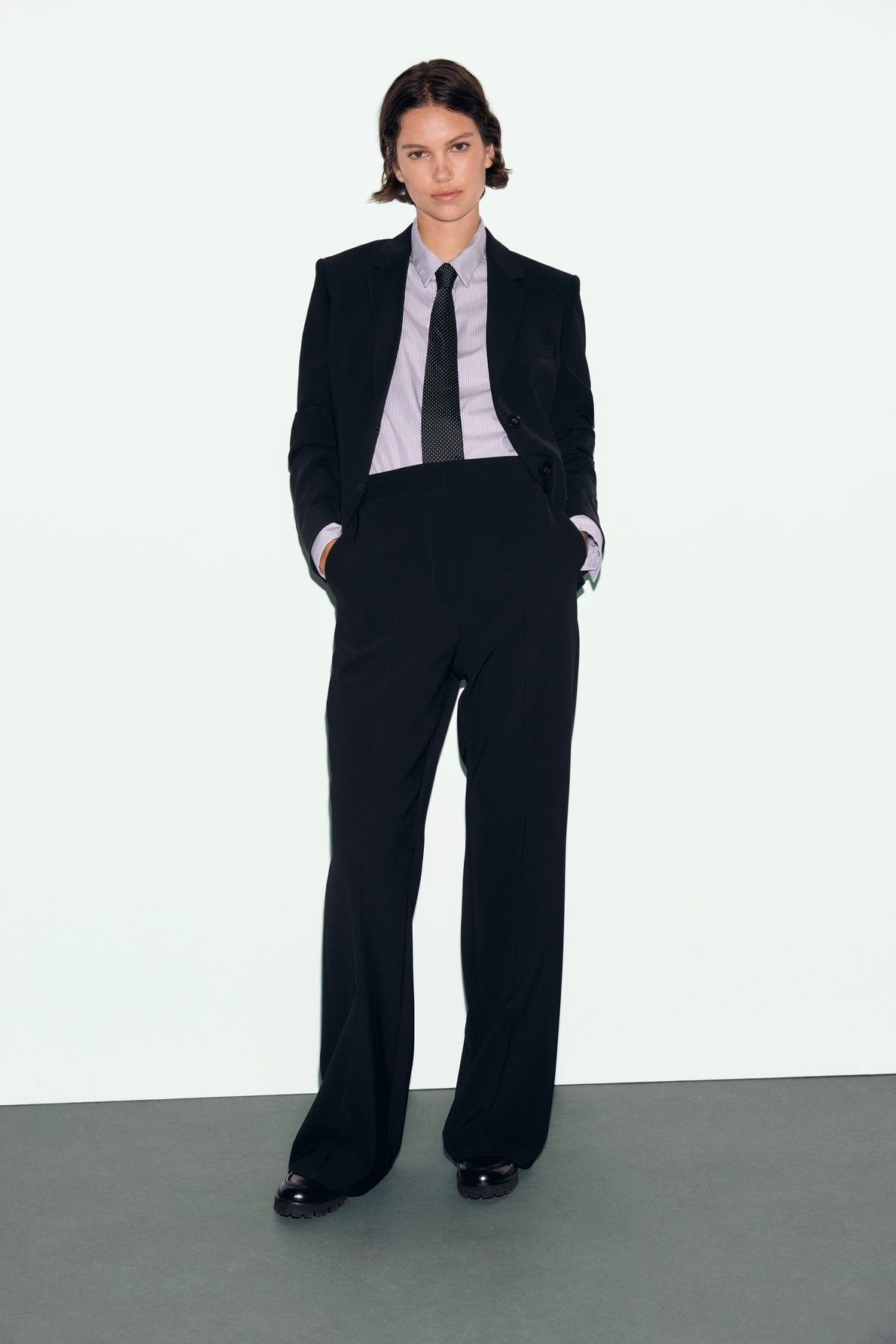 A person stands with hands in pockets, wearing a veste noire de tailleur, white shirt, and black tie, against a plain light background.