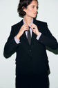 A person in a veste noire adjusts their black tie, looking to the side. They have short, dark hair and are standing against a light-colored background.