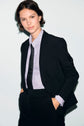 A person with short brown hair wears a veste noire, light striped shirt, and black tie, standing with hands in pockets against a light background.