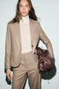 A woman wearing a beige flanelle veste de tailleur and turtleneck stands against a plain background, holding a large brown leather bag under her arm.