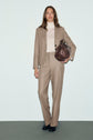Woman wearing a beige flannel veste de tailleur and cream turtleneck, holding a large brown bag under her arm, standing against a plain light background.