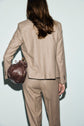 A person with long brown hair, seen from behind, wears a flanelle beige veste de tailleur and carries a dark brown leather handbag on their left shoulder against a plain light background.