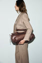 Woman in a flanelle beige veste de tailleur stands in profile, holding a large brown leather duffel bag under her arm against a plain light background.