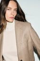 A woman with long brown hair wears a flanelle beige veste de tailleur over a cream turtleneck, looking confidently at the camera against a plain light background.