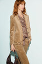 A person with red hair wears a veste tailleur velours beige and a paisley-patterned blouse, holding a dark handbag, standing against a light background.