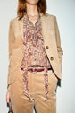 A woman wearing a veste tailleur velours in a chic décontractée style—a tan corduroy suit paired with a brown paisley-patterned blouse and burgundy belt—stands poised against a plain background.