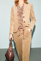 A person wearing a veste tailleur velours beige femme paired with a paisley shirt holds a dark brown handbag. Only the body is visible; the head is cropped out, creating an effortlessly chic décontractée look.