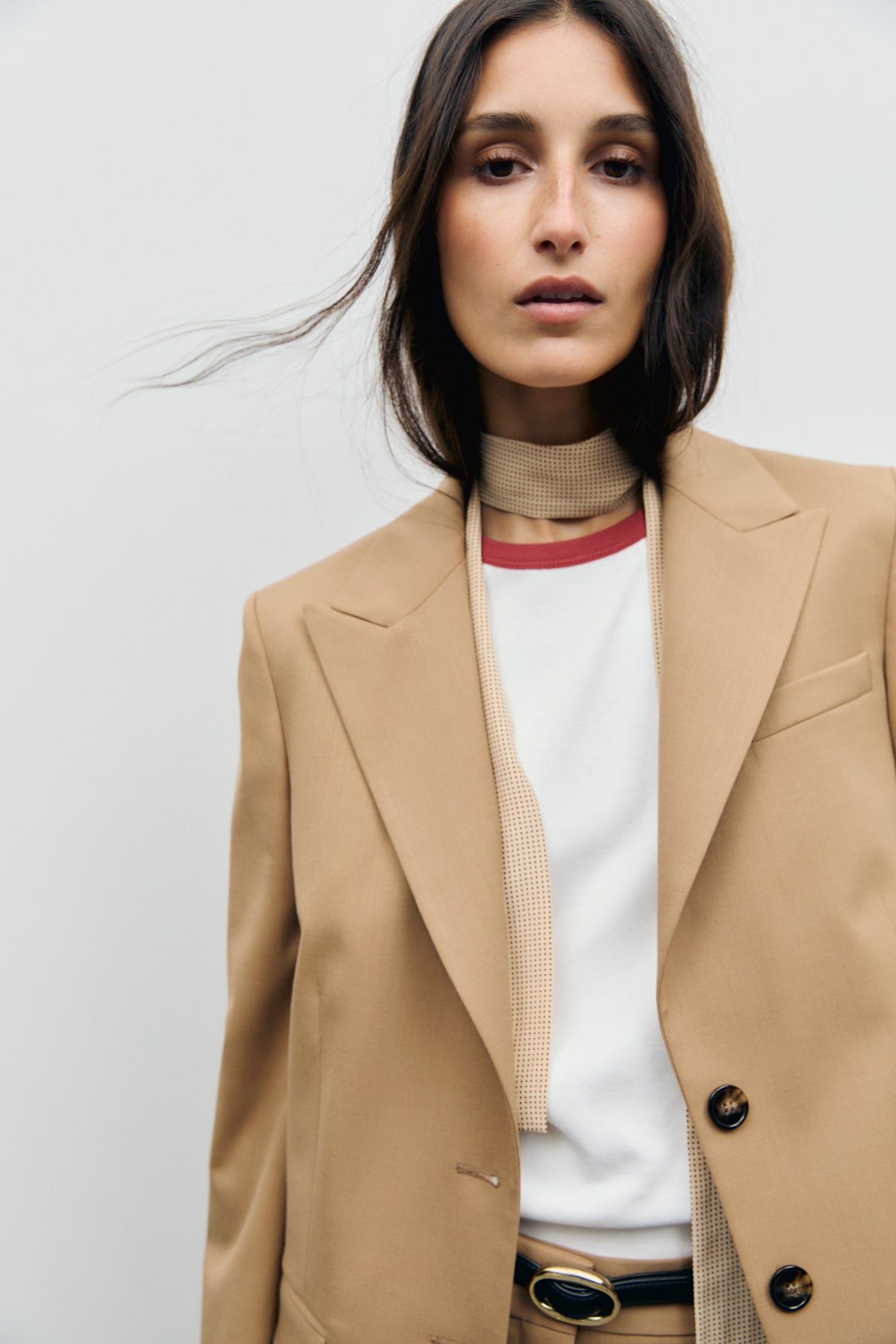 A woman wearing a veste de tailleur camel—styled as a tan blazer—over a white shirt, with a thin beige scarf and black belt, stands against a plain light background, looking into the camera with loose hair framing her face.
