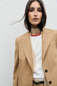 A woman wearing a veste de tailleur camel—styled as a tan blazer—over a white shirt, with a thin beige scarf and black belt, stands against a plain light background, looking into the camera with loose hair framing her face.
