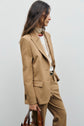 A person wearing a veste de tailleur camel over a white turtleneck and holding a brown bag stands against a plain light background. Their long brown hair is loose, and they are facing sideways.