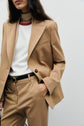 A person wearing a veste de tailleur camel with a white shirt featuring a red neckline, a beige scarf, and a black belt, posing with one hand in their pocket against a plain background.