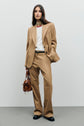 A woman stands against a plain background wearing a veste de tailleur camel, white shirt, and black shoes. She holds a brown handbag in her left hand and has long, straight hair.