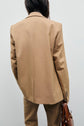 A person wearing a veste de tailleur camel stands facing away from the camera, holding a brown handbag in their left hand against a plain light background.