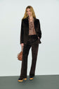 A woman stands in a dark brown veste tailleur velours suit, paisley shirt, platform shoes, and holds a small brown handbag. She has long blonde hair and a neutral expression against a plain background.