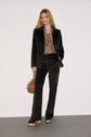 A person in a dark velvet suit—styled like a veste tailleur velours—with a patterned shirt, holding a brown handbag, stands against a plain background. They wear platform shoes and have long, loose hair.