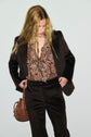 A person with long blonde hair wears a veste tailleur velours in a dark shade, paired with a paisley-patterned blouse and carries a brown handbag while looking down.