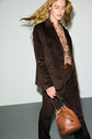 A person with long hair walks while wearing a dark brown veste tailleur velours, a patterned shirt, and carrying a brown leather bucket bag. The background is plain and neutral.