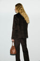 A woman with long blonde hair, wearing a veste tailleur velours dark suit, stands facing away from the camera and holds a small brown leather handbag by her side.