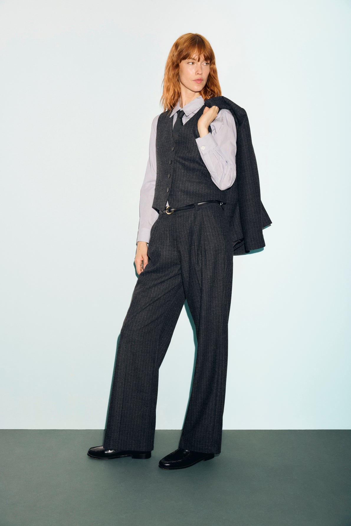 Person with shoulder-length hair wears a flanelle grise pinstripe suit, styled with a gilet de tailleur, and holds the blazer over one shoulder, standing confidently against a plain light background for a look classique chic.