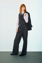 Person with shoulder-length hair wears a flanelle grise pinstripe suit, styled with a gilet de tailleur, and holds the blazer over one shoulder, standing confidently against a plain light background for a look classique chic.