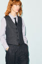 A red-haired model showcases a look classique chic, wearing a tailored gilet de tailleur and matching flanelle grise pinstriped trousers over a light-colored striped shirt with a black polka-dot tie, posing against a light blue background with her hands in her pockets.