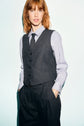 A person with red hair wears a gilet de tailleur in flanelle grise, paired with matching pinstriped trousers, a light blue shirt, and a black tie, embodying a look classique chic as they stand against a plain light background with hands in pockets.
