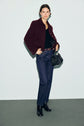 A person stands against a pale background wearing a veste bordeaux in the style of a veste courte femme, paired with a black top, blue jeans, and black boots, holding a black handbag.