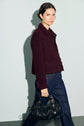 Woman with short dark hair wearing a veste bordeaux courte femme and dark jeans, holding a black woven handbag, standing against a pale grey background.