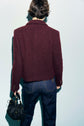 Person with short brown hair wearing a veste tweed bouclé bordeaux and dark jeans is standing with their back to the camera, holding a black quilted handbag in their left hand against a light background.