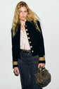 A woman with long blonde hair wears a black veste militaire with gold buttons, a light blouse, patterned pants, and holds an olive green handbag. She stands against a plain light background.