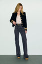 A woman stands against a plain background wearing a black veste col officier with gold buttons, a pale ruffled blouse, patterned high-waisted jeans, a belt, and platform shoes. She has long, wavy hair and looks downward.