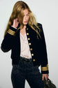 A woman with long blonde hair wears a navy veste militaire with gold buttons and striped cuffs, a white ruffled blouse, and dark patterned jeans. She is looking down and touching her hair.