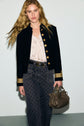 A woman with long blonde hair wears a black veste col officier with gold buttons, a light blouse, patterned high-waisted jeans, and holds a taupe handbag, standing against a plain light background.