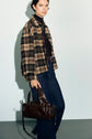 A woman stands against a light background, wearing a surchemise laine imprimé carreaux in brown and black, paired with a dark turtleneck and wide-leg jeans. She holds a dark brown leather handbag by her side and has short dark hair.