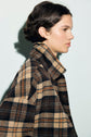 A person with short dark hair wearing a veste courte in brown, black, and beige imprimé carreaux stands in profile against a plain light background.