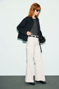 A woman with red hair wears sunglasses, a black jacket styled as a surchemise noire, a black-and-white striped chemise chic, white wide-leg pants, and black shoes, holding a black handbag, standing against a plain light background.