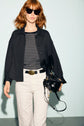 A woman with red hair wears sunglasses, a black jacket styled as a surchemise noire over a striped shirt, white pants, and carries a black handbag against a light blue background.