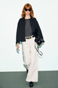 Woman with red hair wears sunglasses, a surchemise noire over a striped top, white wide-leg pants, and black shoes, carrying a black handbag as she walks toward the camera against a plain background.