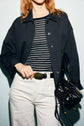 A person with wavy, reddish hair wears a surchemise noire over a black-and-white striped shirt, white pants, and holds a black handbag while adjusting a black belt.