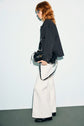A person with red hair, wearing sunglasses, a surchemise noire styled as a black jacket, white wide-leg pants, and black shoes walks holding a black handbag, against a plain light background.