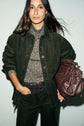 A woman in a dark green surchemise velours côtelé and matching pants, floral-patterned turtleneck, holding a large brown leather bag under her arm with one hand slipped into one of the grandes poches, stands against a light background, looking confidently at the camera.