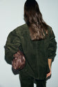 A person with long brown hair, seen from behind, wears a dark green surchemise velours côtelé with grandes poches and carries a brown leather drawstring bag over their left shoulder.