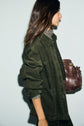 A woman with long brown hair wears a kaki surchemise en velours côtelé with grandes poches over a floral shirt and carries a dark brown leather bag on her shoulder. She is facing sideways against a plain light background.