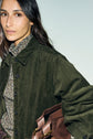 Woman with long dark hair wears a surchemise velours côtelé kaki with grandes poches over a patterned button-up shirt, holding a brown leather bag and looking towards the camera against a light background.