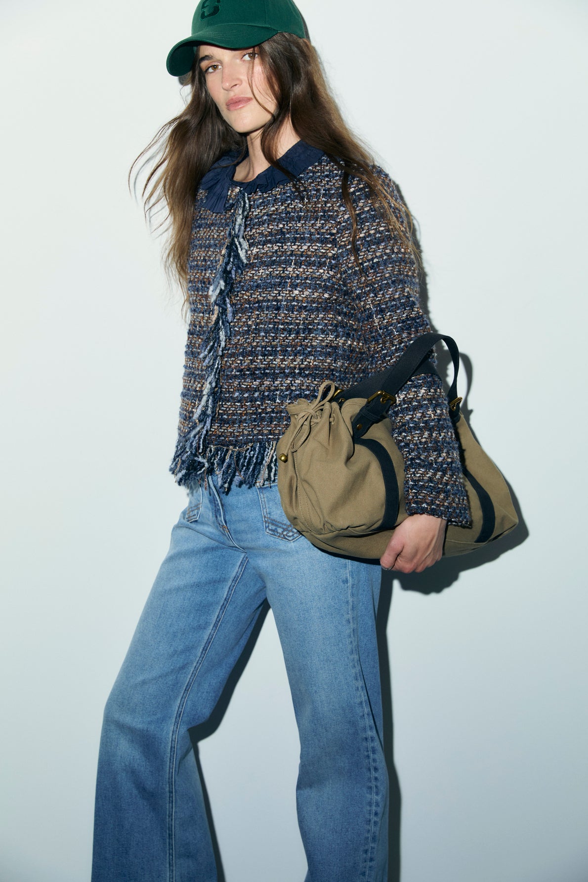 A person wearing a green cap, a veste en tweed courte with tissage multicolore, light blue jeans, and holding a tan bag stands against a plain light background.