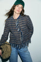 A person wearing a green cap, a veste en tweed—textured blue and brown with fringe—and blue jeans holds a khaki bag, standing against a plain light background.