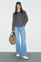 Woman wearing a green cap, veste courte en tweed au tissage multicolore, and flared light blue jeans stands against a plain background, holding a tan bag in one hand and wearing white sneakers.