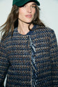 A woman with long brown hair wears a green cap and a veste en tweed courte au tissage multicolore bleu et brun, looking slightly off-camera with a neutral expression against a plain background.