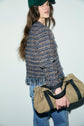 A woman in a green cap, blue veste courte en tweed, and jeans holds a beige bag with black straps. She stands sideways, looking at the camera, against a plain light background.